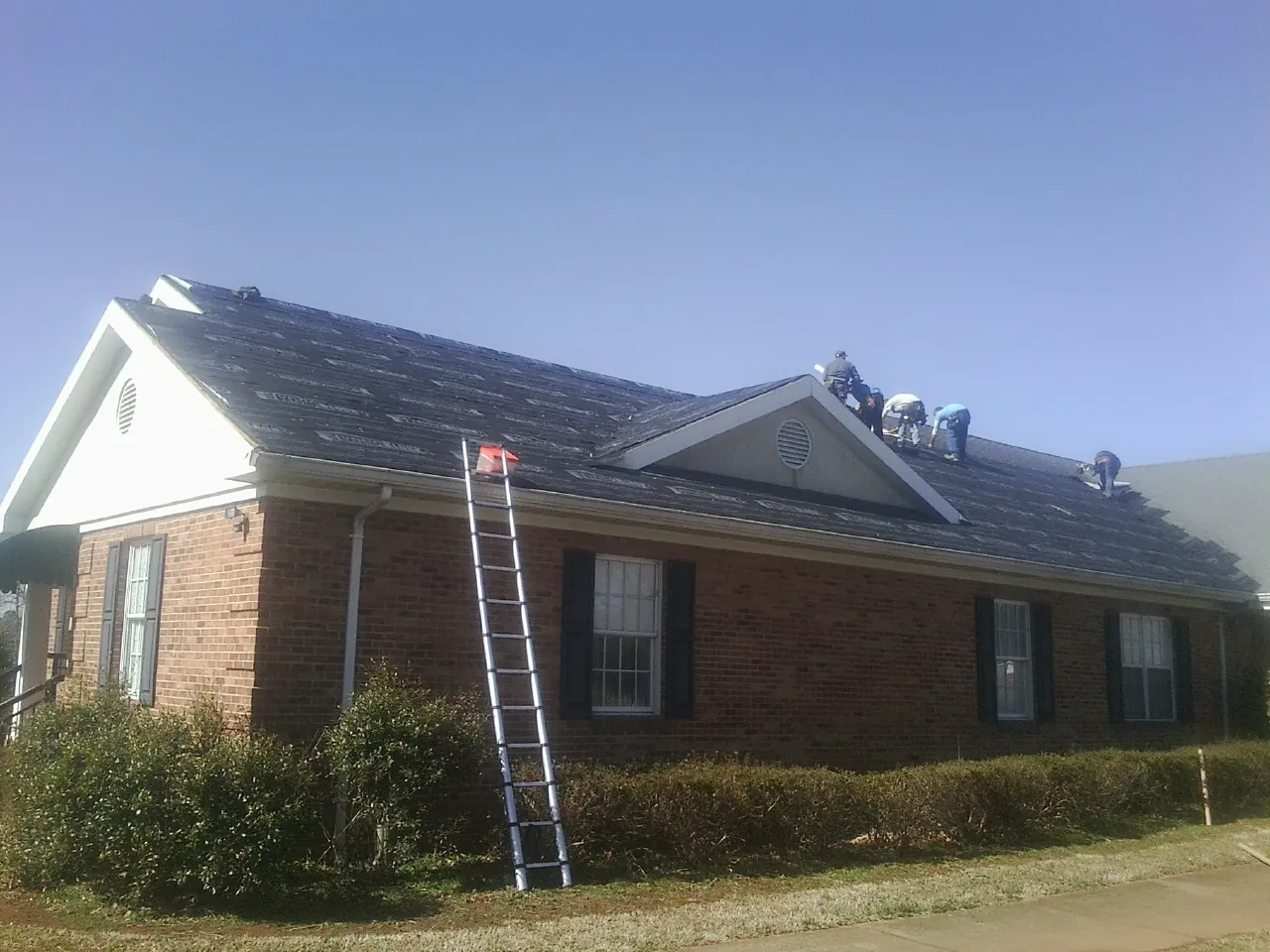 Crew tearing shingles off a brick ranch roof with a ladder set — Emergency Roof Repair work by our crew in Melstone