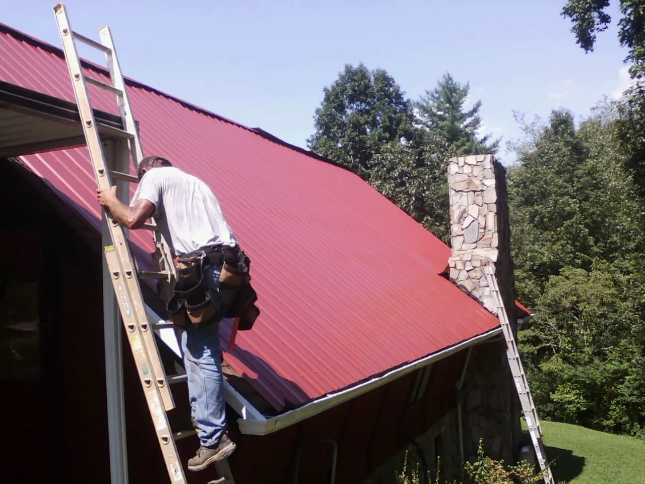 Roofer on a ladder beside a red metal roof and stone chimney — Soffit & Fascia Repair by Roofing Brothers in Melstone